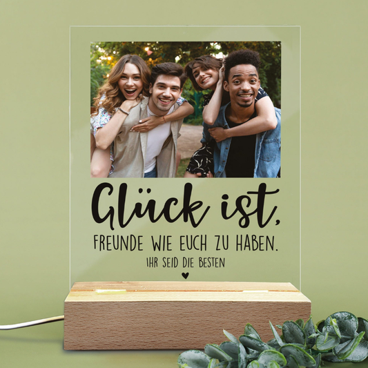 Personalisierte LED-Tischlampe in Herzform mit Foto eines Paares beim Sonnenuntergang und goldener Gravur ‚Ewig Dein‘, platziert auf einem schlichten Holztisch neben einer Geschenkbox und einem Glas Sekt. Warmes Licht erzeugt eine gemütliche Atmosphäre