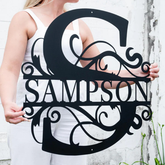 Schwarzes Metall-Monogramm mit dem geteilten Buchstaben ‚M‘ und dem gravierten Familiennamen ‚Schneider‘, montiert an einer rustikalen Holzverkleidung. Umgeben von Topfpflanzen und einer LED-Lichterkette, die abends warmes Licht auf das Design wirft