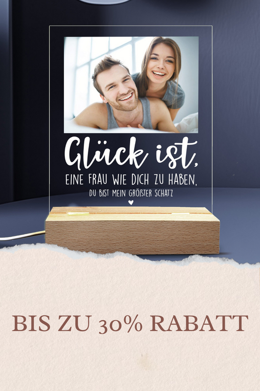 Personalisierte LED-Tischlampe in Herzform mit Foto eines Paares beim Sonnenuntergang und goldener Gravur ‚Ewig Dein‘, platziert auf einem schlichten Holztisch neben einer Geschenkbox und einem Glas Sekt. Warmes Licht erzeugt eine gemütliche Atmosphäre